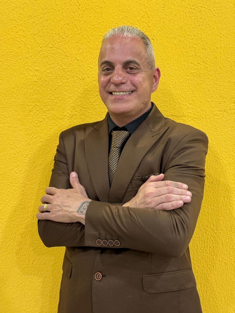 Dinho do Canella sorrindo, de terno marrom, diante de um fundo amarelo